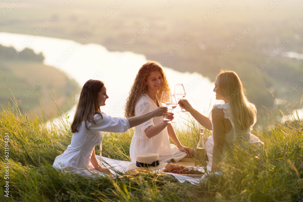 Young and happy women who enjoys amazing scenery sunset at summer picnic. They clinking glasses with wine at sunset.