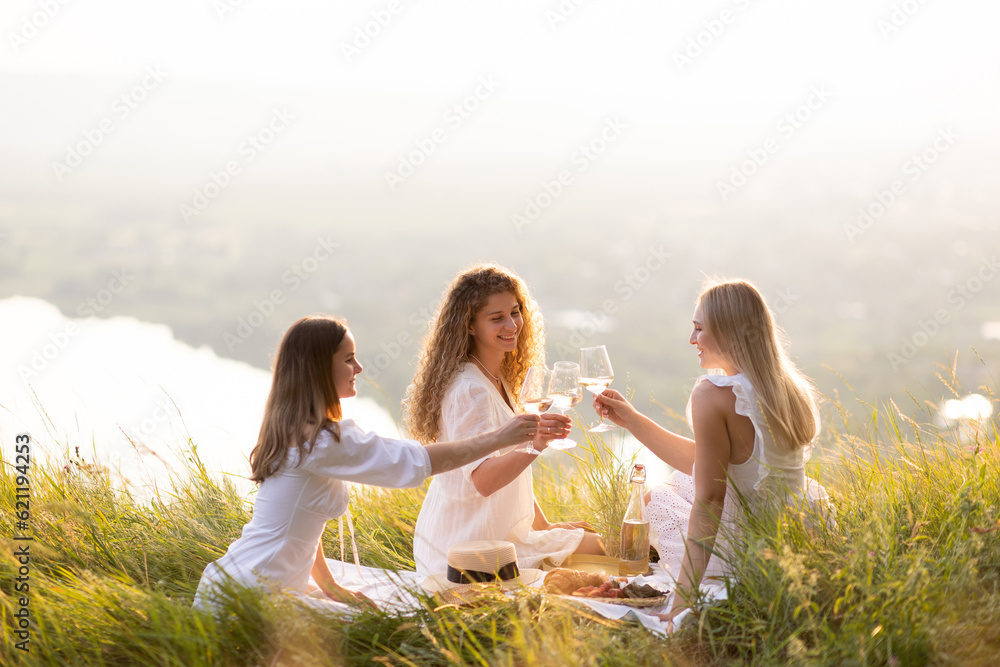 Young girlfriends enjoying picnic on the hill at sunset. Happy friends drinking wine.