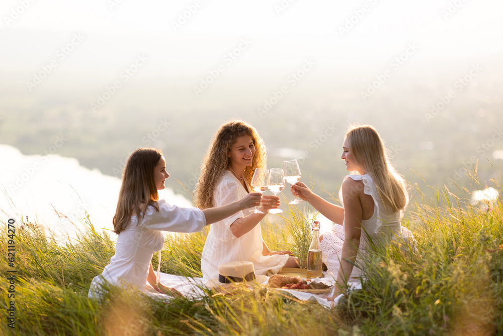 Young women enjoying summer picnic at sunset on the hill. They clinking glasses of wine to celebrate their free time.