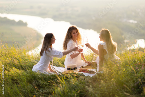 Obraz na płótnie Happy girlfriends sitting at summer picnic with wine and watching sunset at a mountain, while making a toast with wine during golden hour. w salonie