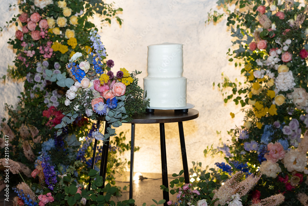 White wedding cake in the evening. Cake on the background of an arch of fresh flowers.