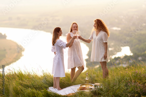 Company of young female friends having fun, raising glasses with wine and enjoy beautiful sunset at summer picnic on the hill.