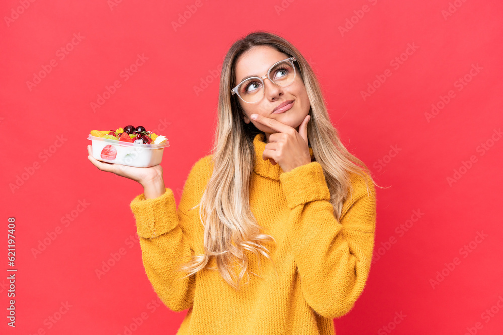 Young pretty Uruguayan woman holding a bowl of fruit isolated on red background having doubts while looking up Young pretty Uruguayan woman holding a bowl of fruit isolated on red background having doubts while looking up