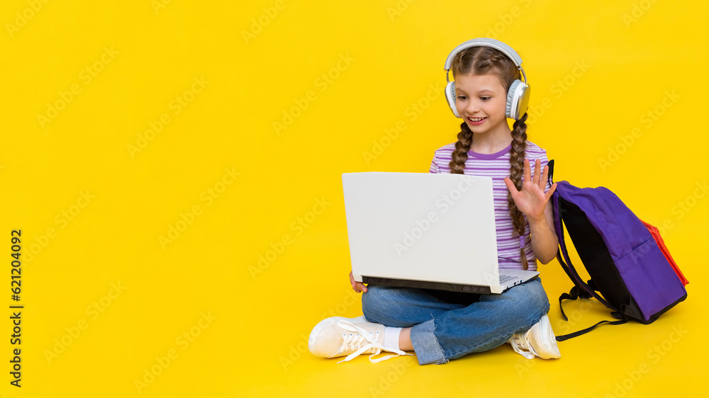 The child is engaged in an online lesson with a laptop. A young girl is sitting cross-legged on the floor with a backpack and headphones. Yellow isolated background. Copy space. The child is engaged in an online lesson with a laptop. A young girl is sitting cross-legged on the floor with a backpack and headphones. Yellow isolated background. Copy space.