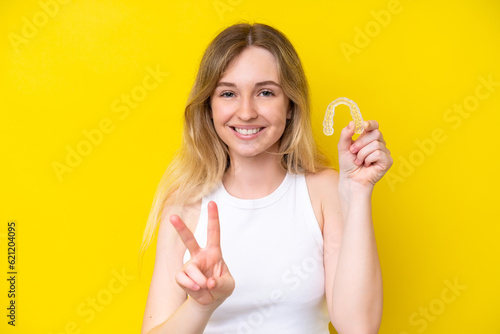 Blonde English young girl holding invisible braces isolated on yellow background smiling and showing victory sign