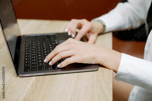 Hands of young freelancer typing on keyboard of laptop. Lady in white shirt working online sitting on couch at table.