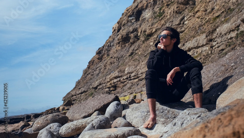 Foto Thoughtful, attractive young man is sitting on large sea stones, On a summer day, A guy is relaxing on the seashore during a bright day