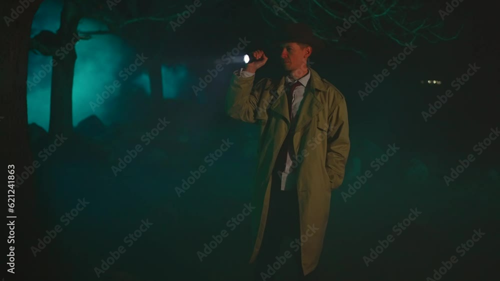Old fashioned detective man in a hat with a flashlight in his hands in a dark forest is searching for clues Old fashioned detective man in a hat with a flashlight in his hands in a dark forest is searching for clues