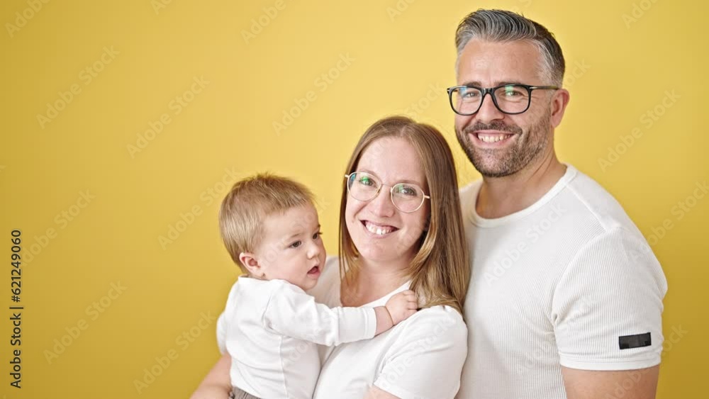 Family of mother, father and baby smiling together looking a the camera over isolated yellow background Family of mother, father and baby smiling together looking a the camera over isolated yellow background