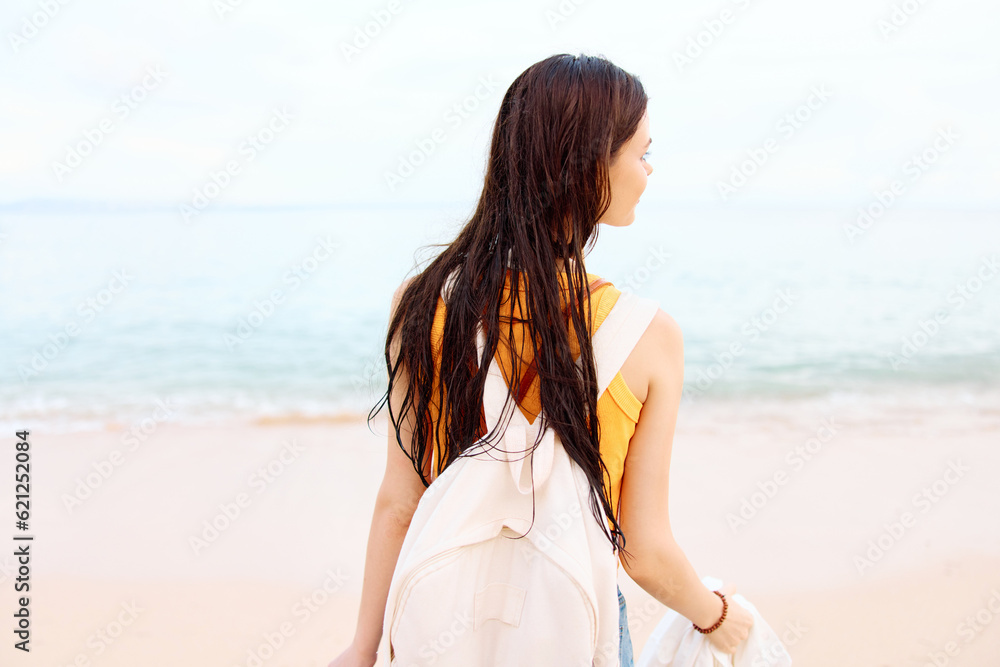 Young woman smile with teeth after swimming in the ocean with a backpack in wet clothes walking along the beach, summer vacation on the island by the ocean in Bali sunset