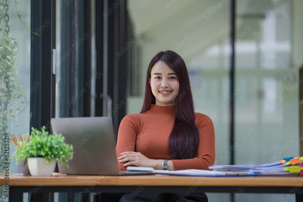 Portrait of a young businesswoman using a laptop, smiling, and looking at the camera while sitting at an office desk in a modern office. Portrait of a young businesswoman using a laptop, smiling, and looking at the camera while sitting at an office desk in a modern office.