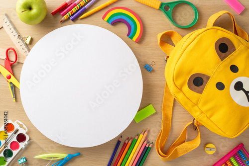 Early education concept. Top-view shot of vivid supplies, painting materials, scissors, ruler, magnifier, plasticine, playful bear-shaped backpack on wooden tabletop with empty circle for text or ad
