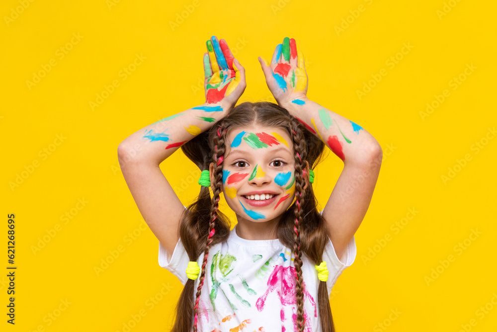 Portrait of a girl with a face painted with multicolored paints. Children's creativity. The child shows his ears above his head. Yellow isolated background. Portrait of a girl with a face painted with multicolored paints. Children's creativity. The child shows his ears above his head. Yellow isolated background.