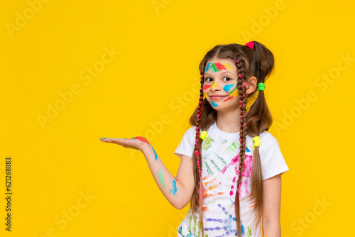 A child smeared with paint. A little girl, painted with multicolored paints, holds your advertisement in the palm of her hand. Children's creativity. Yellow isolated background. Copy space. Banner.