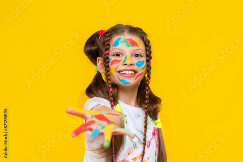 Drawing and creativity for children. A little girl shows her palms painted with multicolored paints. A child smeared in paint. Yellow isolated background.