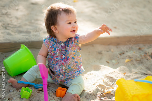 Adorable 9 months old baby playing outdoors - lifestyle portrait of mixed ethnicity Asian Caucasian baby girl playing with block toys happy and carefree at playground sitting on sand