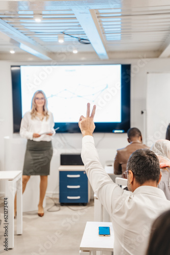 Senior businessman raising a hand to ask a question while his blonde, female, business trainer explaining something