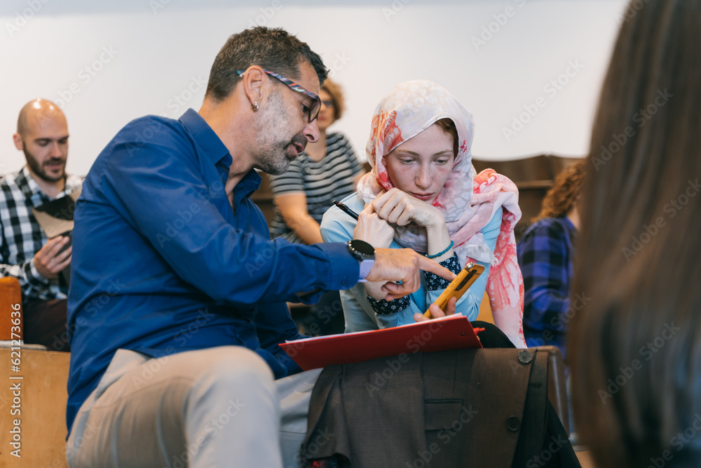 Obraz premium Middle-aged, experienced, male employee helping his muslim junior coworker. Male employee calculating on phone, he is helping his female colleague
