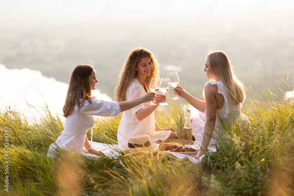 Group of smiling female best friends sitting together on blanket enjoying at picnic in the park at sunset.