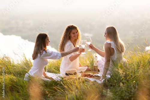 Obraz na płótnie Group of smiling female best friends sitting together on blanket enjoying at picnic in the park at sunset. w salonie
