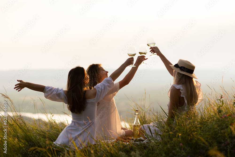 Three young beautiful happy woman is resting at picnic at sunset. Girlfriends enjoy sun day evening on beautiful green meadow. Vacation, picnic, friendship or holiday concept