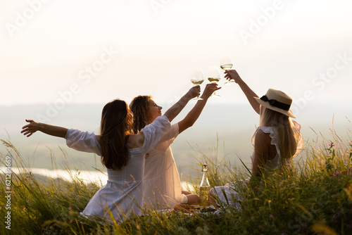 Obraz na płótnie Three young beautiful happy woman is resting at picnic at sunset. Girlfriends enjoy sun day evening on beautiful green meadow. Vacation, picnic, friendship or holiday concept w salonie