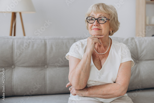 Smiling senior blonde haired woman looking at camera, happy old lady in glasses posing at home indoor, positive single senior retired female sitting on sofa in living room headshot portrait © anatoliycherkas
