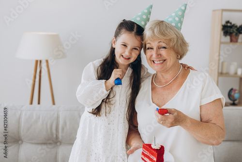 Happy smiling grandmother receives birthday greetings from granddaughter  little girl and senior woman blow out candle on cake and celebrate birthday together.