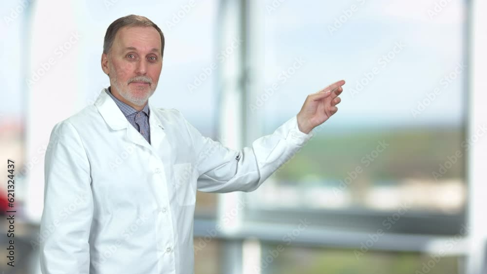 Pensive senior male doctor in white coat touching his chin and showing copy space with his hand. Blurred indoor window in the background. Pensive senior male doctor in white coat touching his chin and showing copy space with his hand. Blurred indoor window in the background.