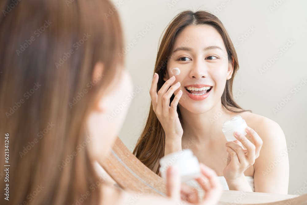 Facial beauty skin care, smile of pretty asian young woman in bathrobe looking at mirror, hand applying moisturizer lotion on her face, holding jar of skin cream before makeup cosmetic routine at home Facial beauty skin care, smile of pretty asian young woman in bathrobe looking at mirror, hand applying moisturizer lotion on her face, holding jar of skin cream before makeup cosmetic routine at home
