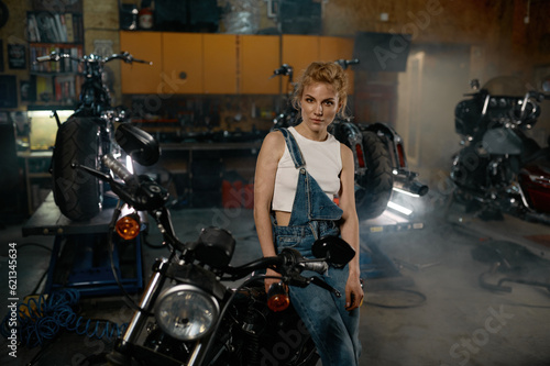 Young woman in denim overalls looking at camera posing at motorcycle garage