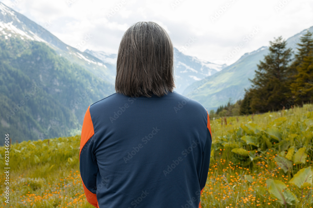 Long-haired man with gray hair sitting with his back in a flower meadow against the backdrop of green lawn and mountains
