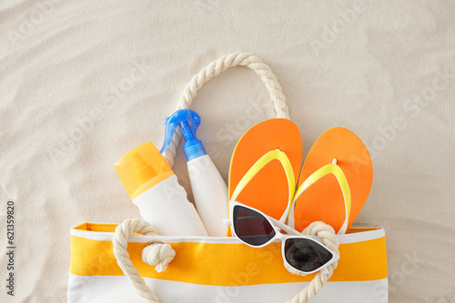 Beach relaxation concept. Top view composition of sunblock cream bottles, beach handbag, orange flip-flops and eyewear on sand background
