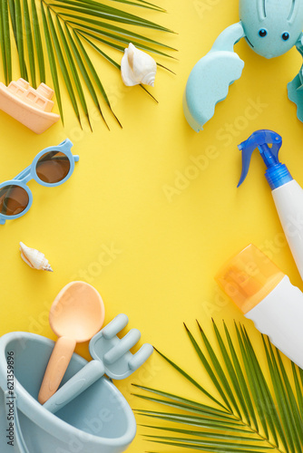 Summer beach getaway for children concept. Top view vertical photo of sunscreen bottles, beach toys, eyeglasses, palm leaves, seashells on yellow background with blank space for advert or text