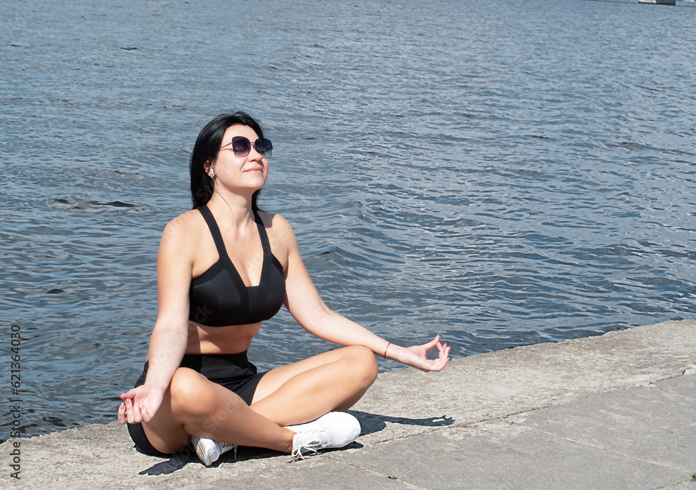 Yoga concept. A beautiful girl in a lotus position sits on the embankment by the river. Meditate with closed eyes.