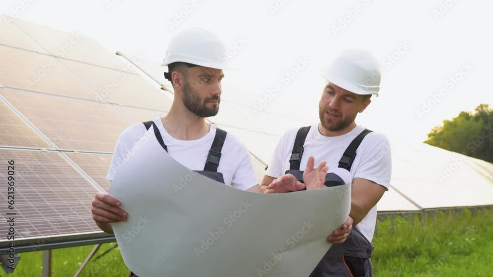 Two workers engineers talking with each other while holding a blue print with a large solar panel in background Two workers engineers talking with each other while holding a blue print with a large solar panel in background