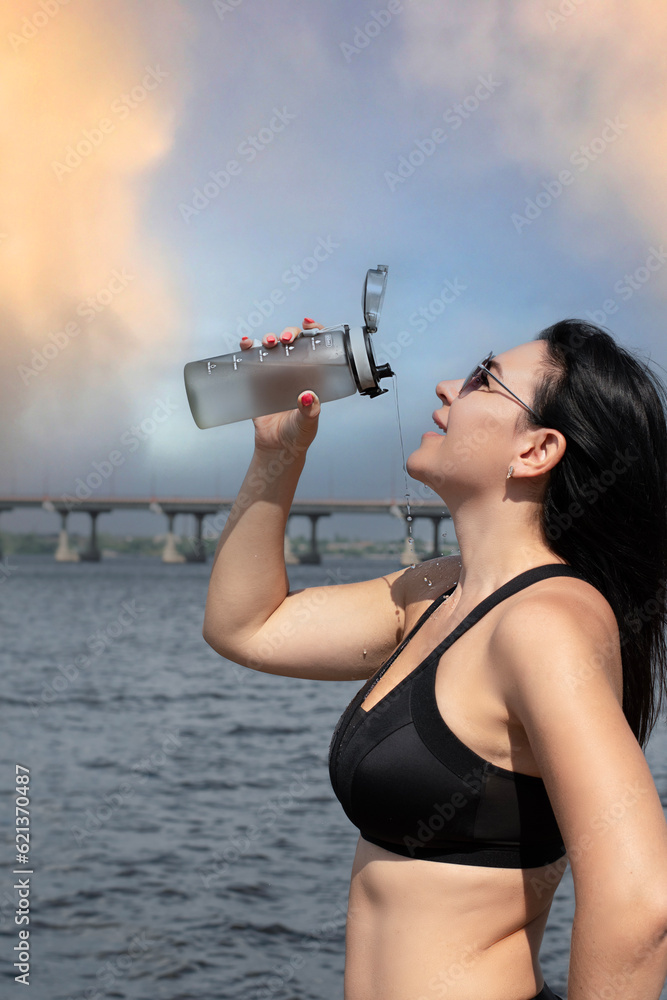 A beautiful and slender girl in sunglasses pours water over herself from a sports bottle against the sky with a rainbow. Sport concept. Close-up. A beautiful and slender girl in sunglasses pours water over herself from a sports bottle against the sky with a rainbow. Sport concept. Close-up.