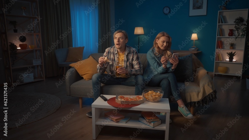 A man with a glass of beer, pizza, watching a sports game. A woman sits next to the phone, scrolls through social networks, ignoring the man, not interested in his hobbies. Various family interests. A man with a glass of beer, pizza, watching a sports game. A woman sits next to the phone, scrolls through social networks, ignoring the man, not interested in his hobbies. Various family interests.