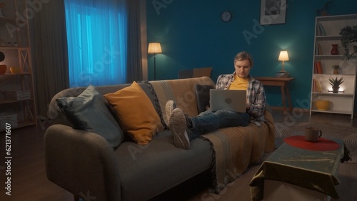 A young man is lying on the couch in the living room with a laptop on his lap. A man with a serious face is working or playing on a laptop, watching a movie, photo, video.