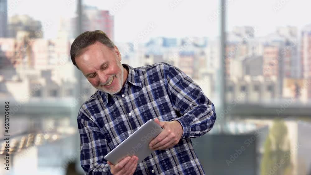 Aged mature man standing indoors playing a video game on digital tablet. Blurred windows in the background. Aged mature man standing indoors playing a video game on digital tablet. Blurred windows in the background.