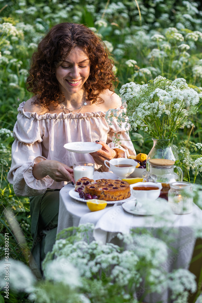 Beautiful curly haired young woman in straw hat drinking herbal tea in the garden, enjoying life, smiling. Simple pleasures, elegance. Fancy white porcelain cup, homemade cherry pie