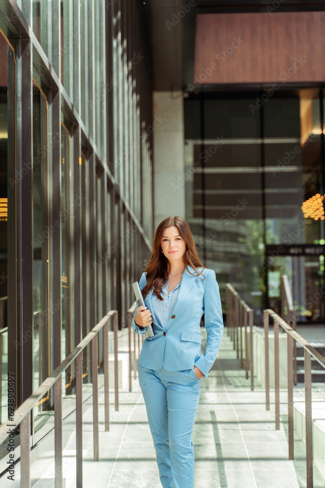 Beautiful businesswoman wearing stylish business suit, holding laptop and looking at camera outdoors Beautiful businesswoman wearing stylish business suit, holding laptop and looking at camera outdoors