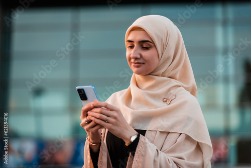 Canvas-taulu Portrait of a happy Muslim business woman in hijab using a phone near the office building
