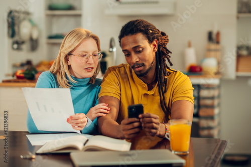 A multicultural couple is planning a budget together while holding a phone and paperwork at home. photo