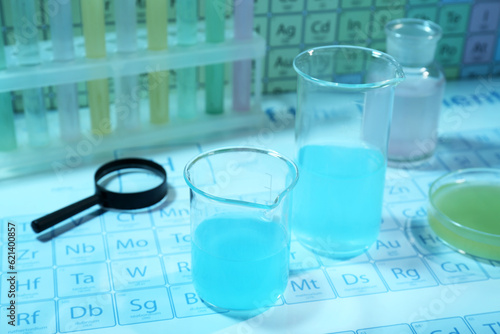 Beakers with liquid, different laboratory glassware and magnifying glass on periodic table of chemical elements. Light blue tone effect