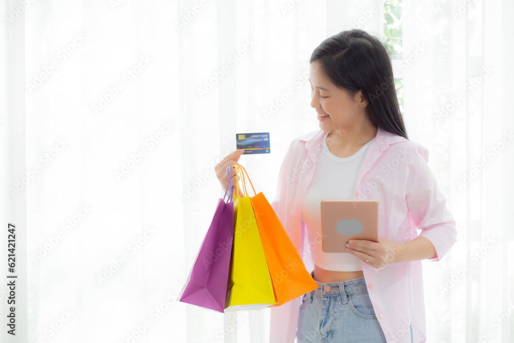 Young asian woman standing using digital tablet shopping online with credit card while holding paper bag, female paying with transaction financial, purchase and payment, business concept. Young asian woman standing using digital tablet shopping online with credit card while holding paper bag, female paying with transaction financial, purchase and payment, business concept.