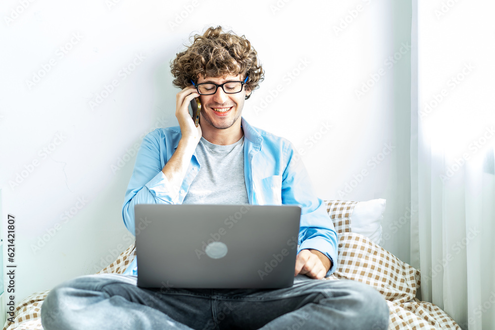 Caucasian Freelancer Guy Talking On Phone Using Laptop Computer Communicating Online Working on the bed in the room, Distance Work And Mobile Communication Concept.