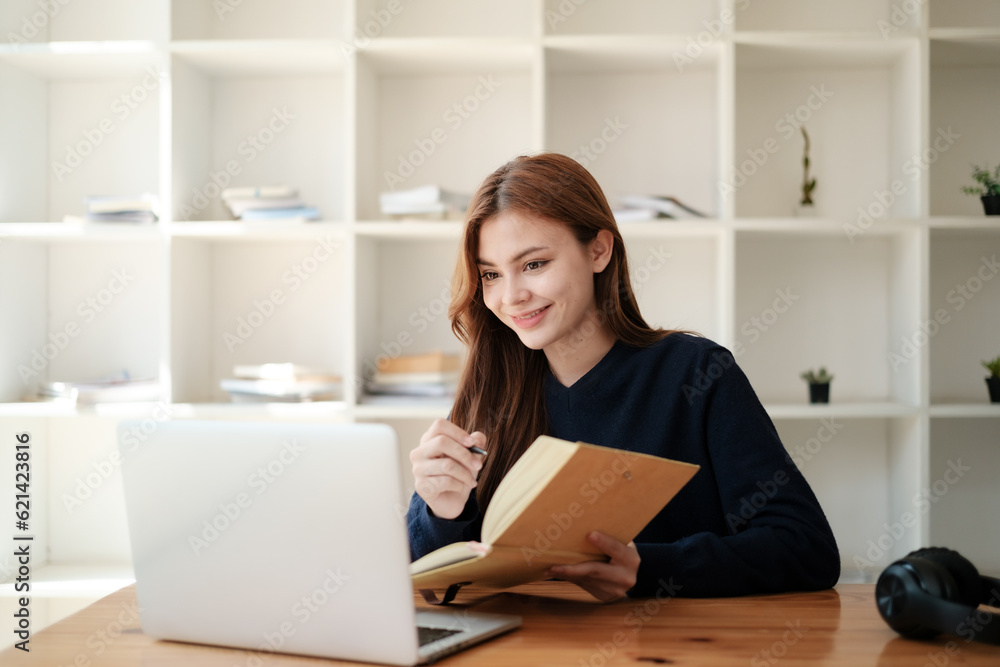 A beautiful caucasian female student is studying in college remotely. She is sitting at a table at home with a laptop and a notepad and concentrated is watching a video conference lesson A beautiful caucasian female student is studying in college remotely. She is sitting at a table at home with a laptop and a notepad and concentrated is watching a video conference lesson