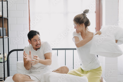 Funny asian couple angry wife hit husband with pillow in white bedroom.