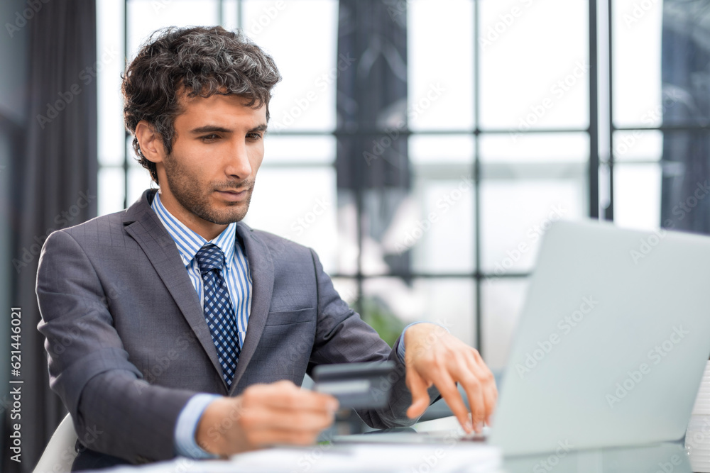 Smiling man sitting in office and pays by credit card with his laptop. Smiling man sitting in office and pays by credit card with his laptop.